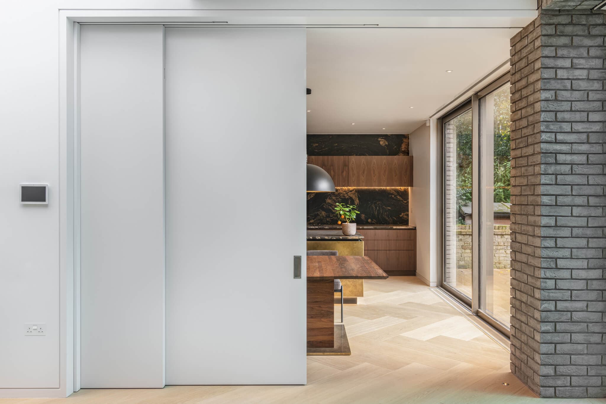 A set of half open white sliding telescopic doors leading into a wood and marble kitchen with wooden flooring and a dark grey brick wall to the right.