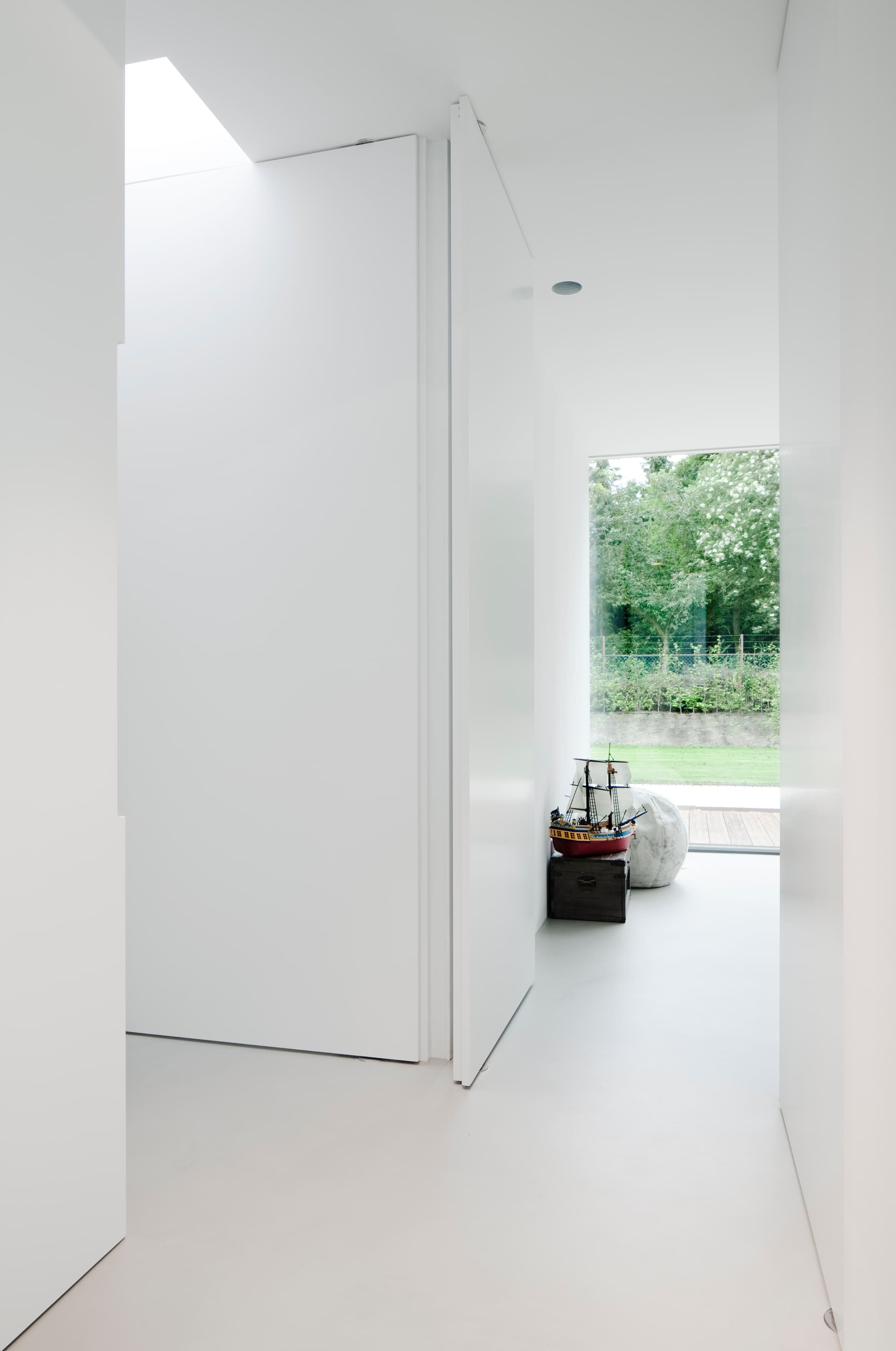 A white minimal interior with closed pivoting door and opening into a bedroom.