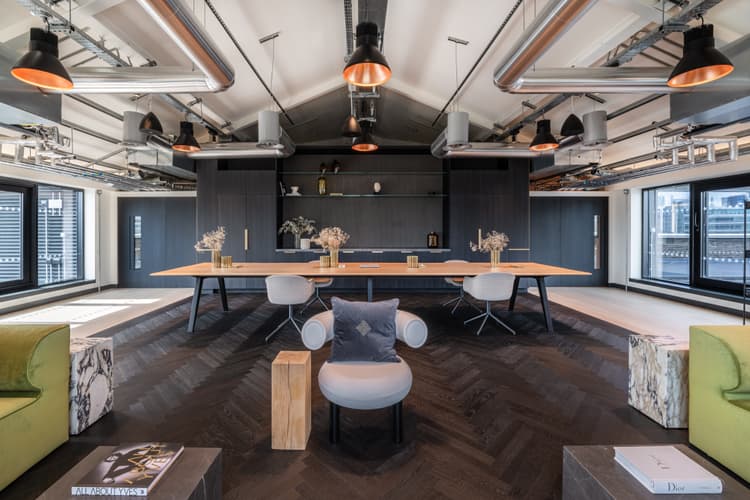 A large office space lounge and dining area with dark flooring, high industrial ceiling, green sofas and dark wood cabinetry and doors.